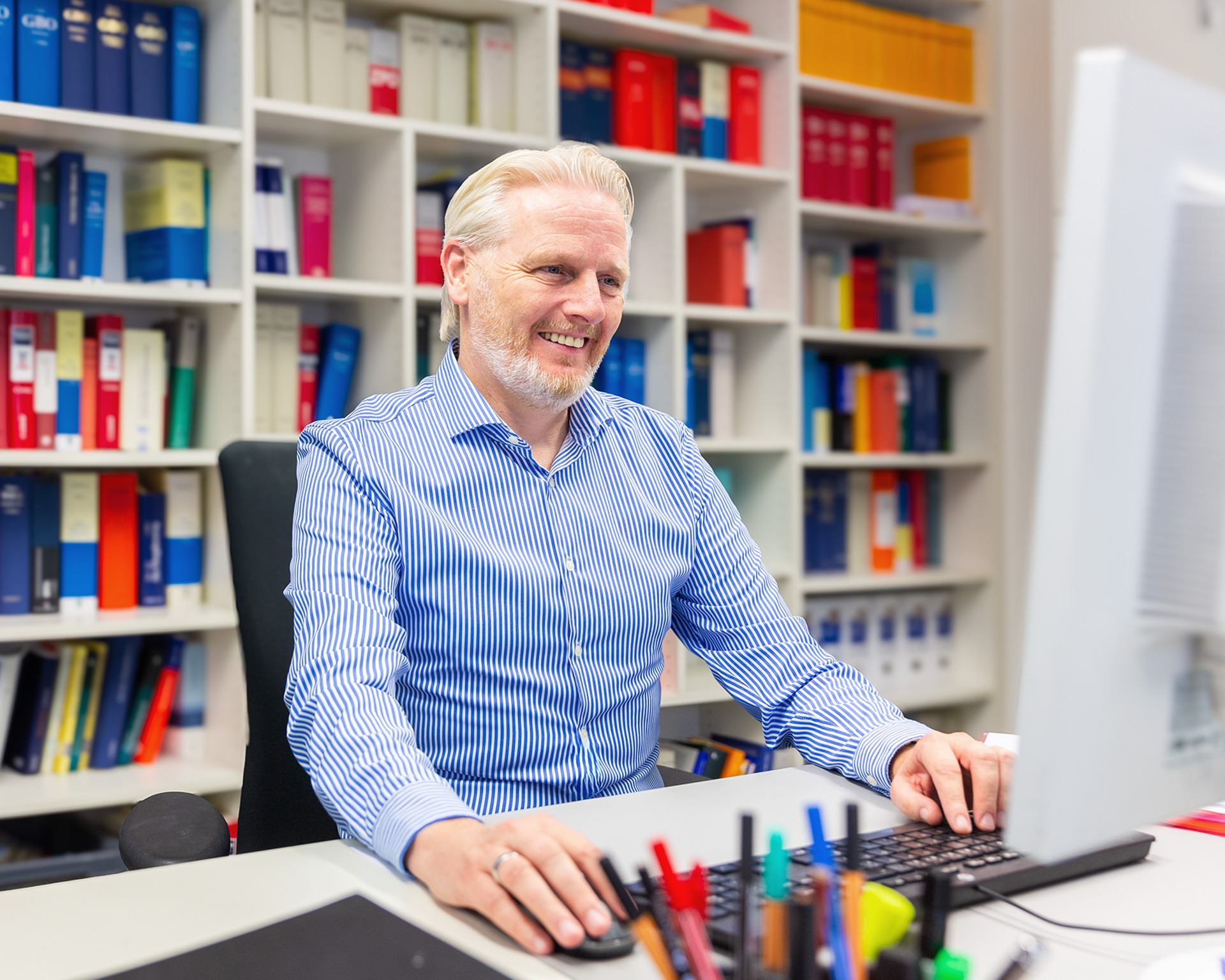 Der Mitarbeitende Robert Müller sitzt an einem Computer, hinter ihm ist ein großes Bücherregal zu sehen